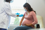 Wearing protective masks because of COVID-19, a mid adult pregnant woman watches as the female technician administers a booster shot