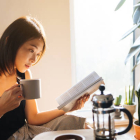 Beautiful young woman reading a book while drinking coffee, enjoying the morning sunlight